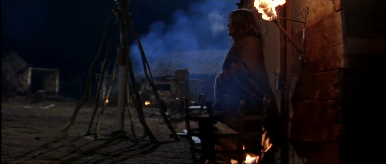 Ravenous (1999) movie still: medium — A nighttime medieval encampment with wooden structures, a tripod frame, and rising smoke. A woman wrapped in a…