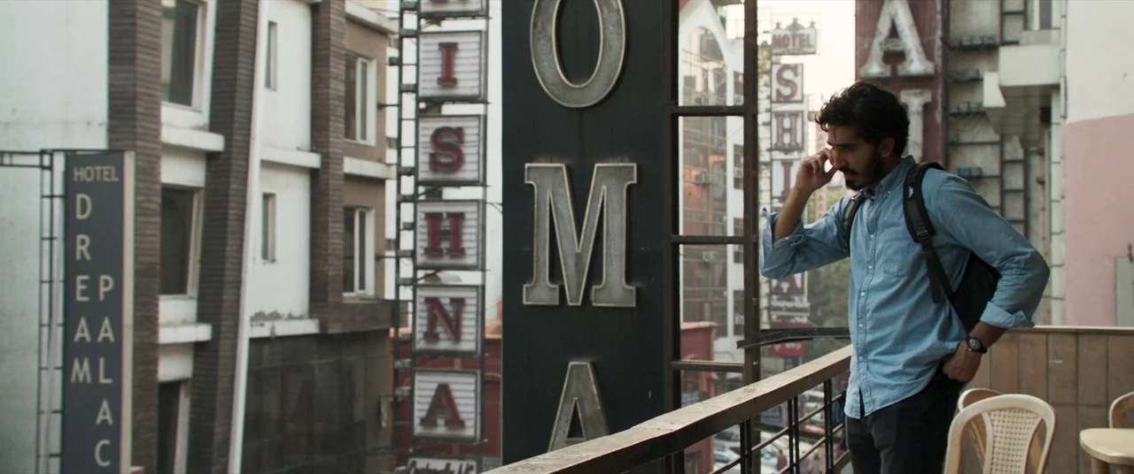 The Wedding Guest (2018) movie still: medium — An urban building exterior features stacked hotel signs with text against a concrete facade. A man in…