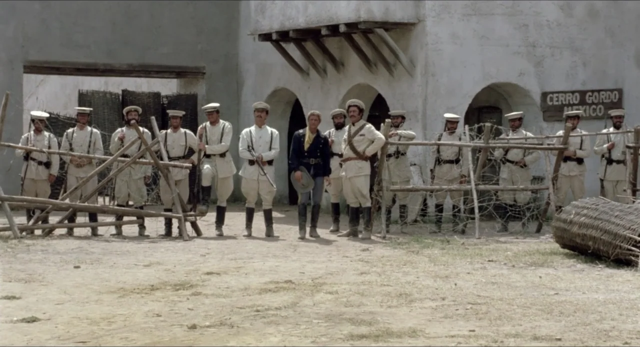 A Stranger in Town (1943) movie still: wide — A dusty military encampment features white stucco arches and a makeshift wire fence barrier. A group…