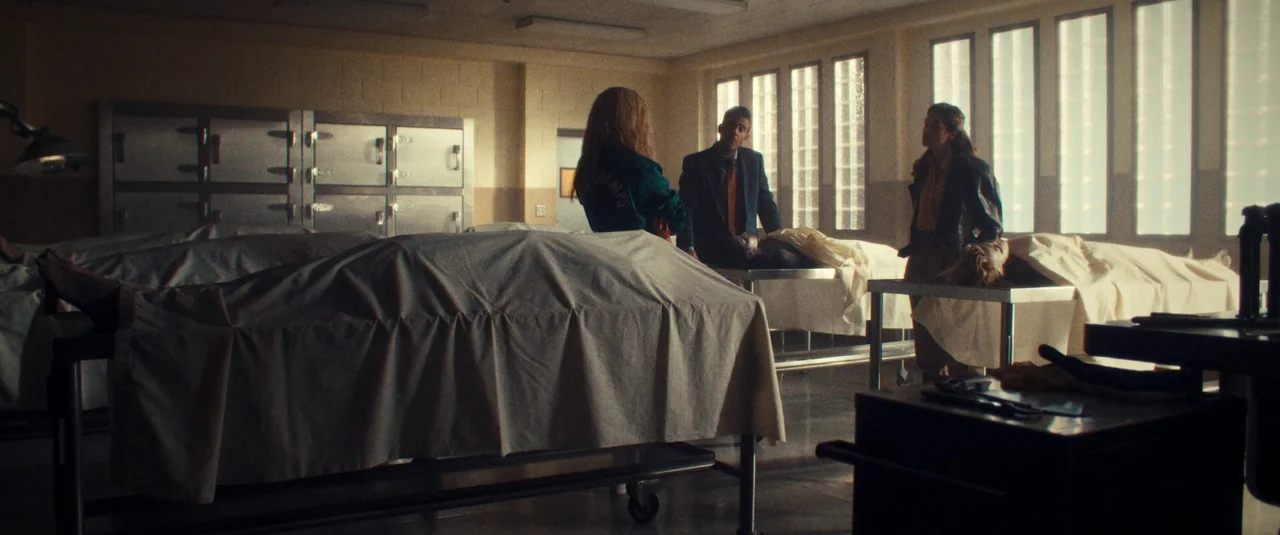 MaXXXine (2024) movie still: wide — A sterile room features rows of metal storage lockers and several body-covered gurneys. Three people stand among…