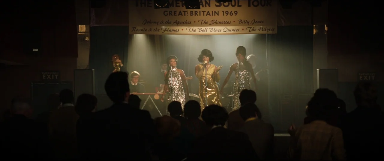 Rocketman (2019) movie still: wide — A dimly lit indoor music venue with a banner referencing a 1969 tour. Three female singers in sparkly gowns…