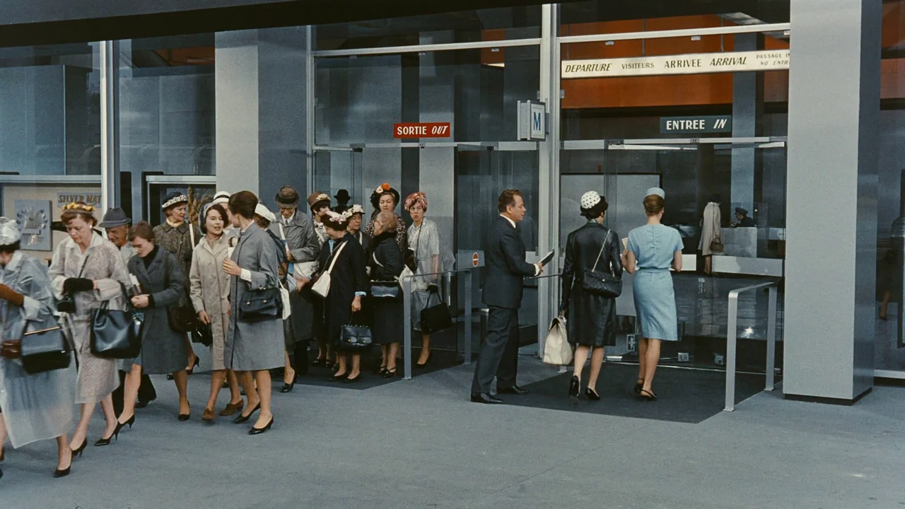 Playtime (1967) movie still: wide — A modern 1960s airport entrance with floor-to-ceiling glass and metal framing. Passengers with handbags and…
