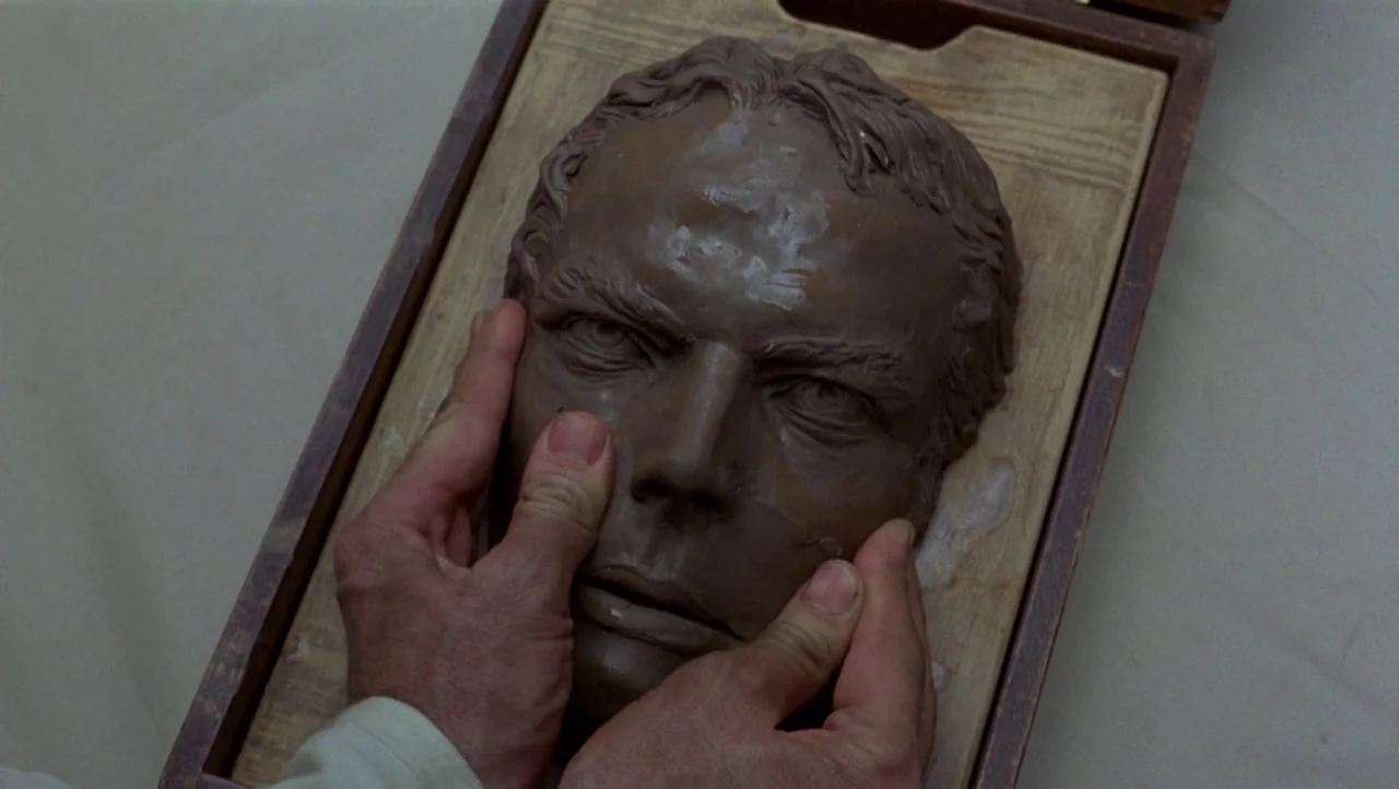 Mountains of the Moon (1990) movie still: closeup — A sculpted clay portrait of a man's face sits inside a wooden frame. Two hands press into the…