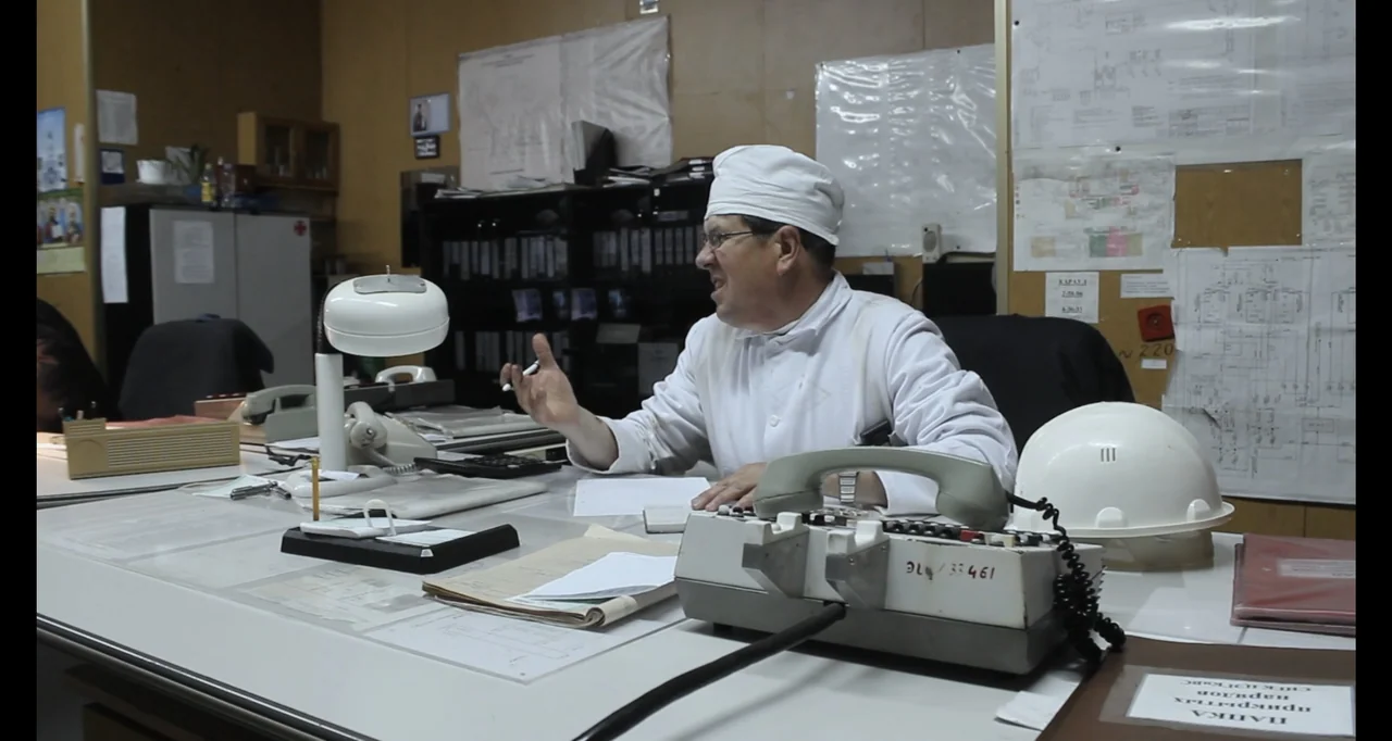 Chernobyl's Café (2016) movie still: medium — An industrial office features papers, technical diagrams on the wall, and multiple telephones. A…