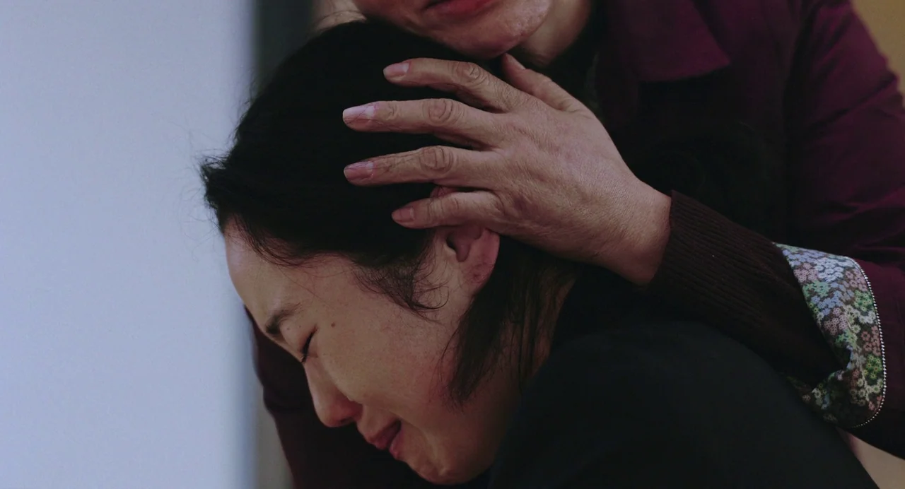 Return To Seoul (2022) movie still: closeup — A close-up view of two individuals inside an indistinct indoor environment. An older person gently…