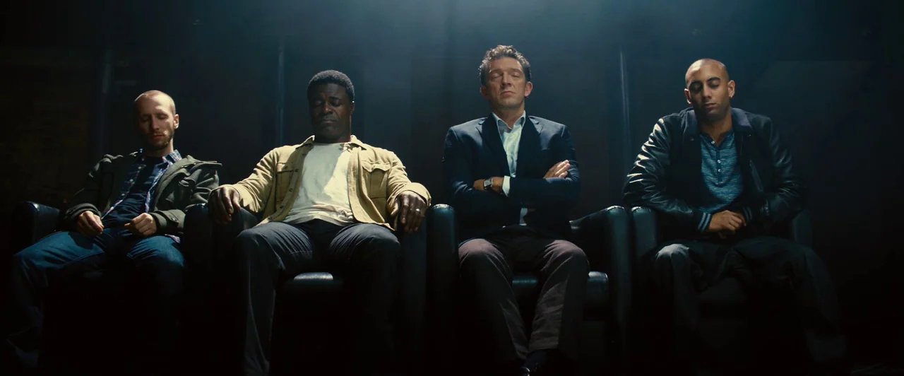 Trance (2013) movie still: medium — Four men sit in leather chairs arranged in a line against a dark wall. Each man sits with his eyes closed under…