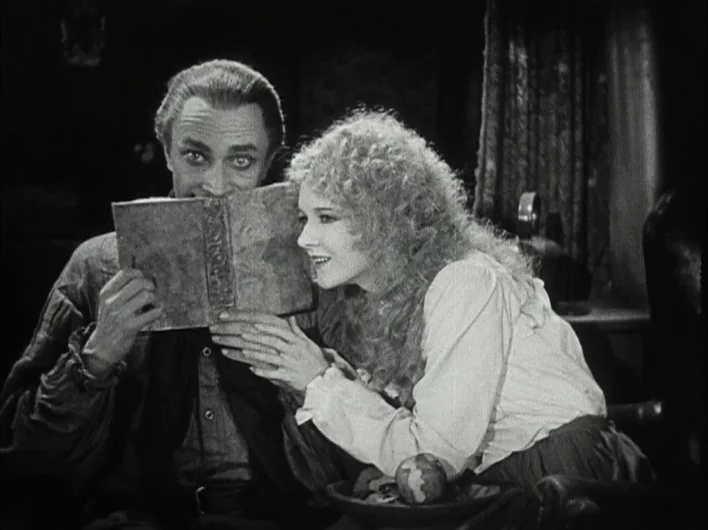 The Man Who Laughs (1928) movie still: medium — A man and woman hold a book together in a dimly lit, dark space. The two figures sit closely while…