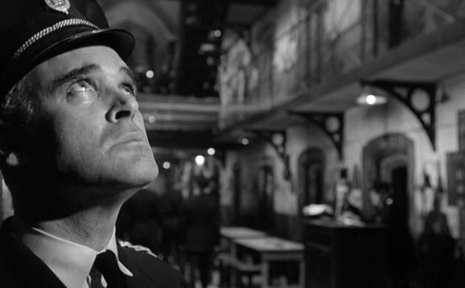 The Criminal (1960) movie still: closeup — A multi-level prison block features arched walkways and iron railings in the background. A guard in…