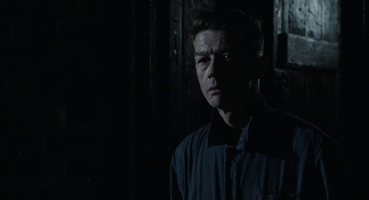 1984 (1984) movie still: medium — A dark space with textured walls and a wooden door frame in the background. A man stands in low light looking…
