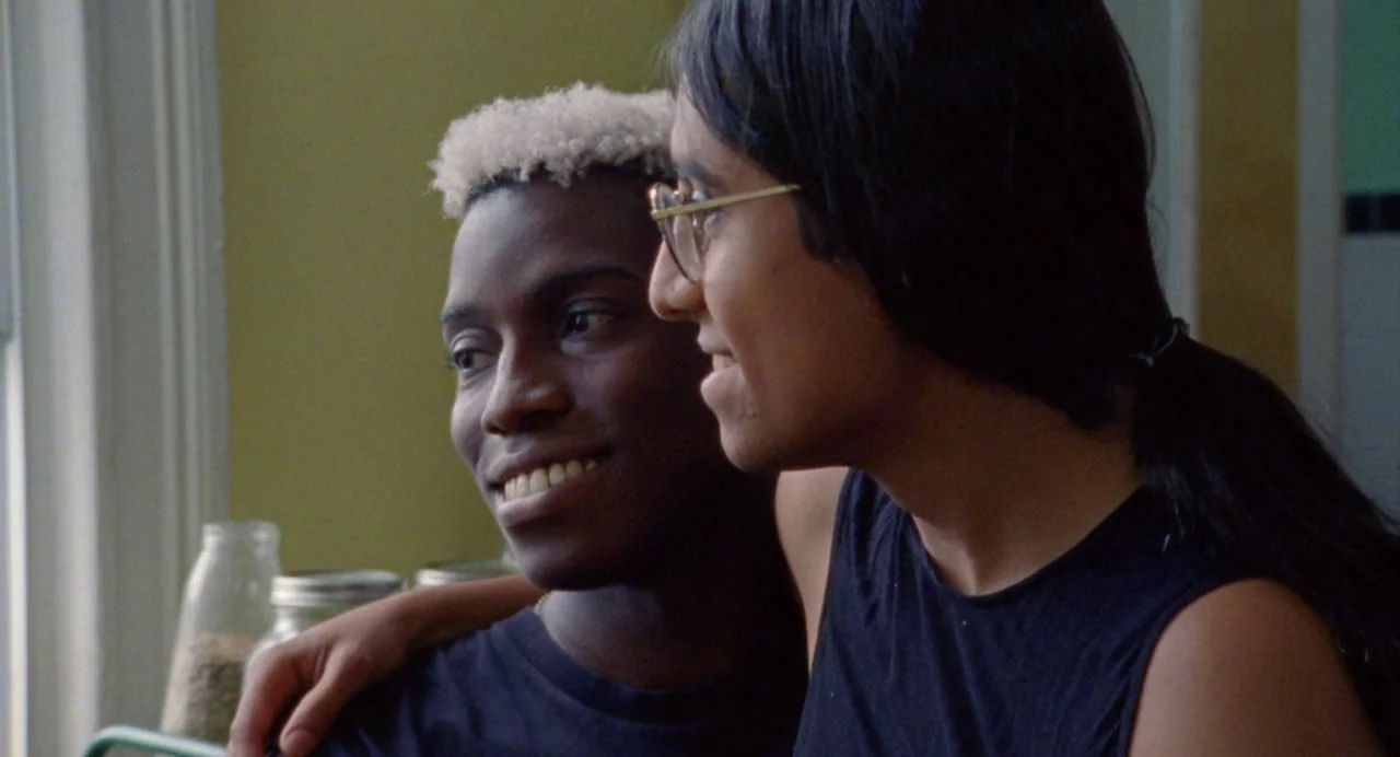 So Pretty (2019) movie still: medium — Two men stand together indoors against a plain yellowish wall in daylight. One man has his arm around the…