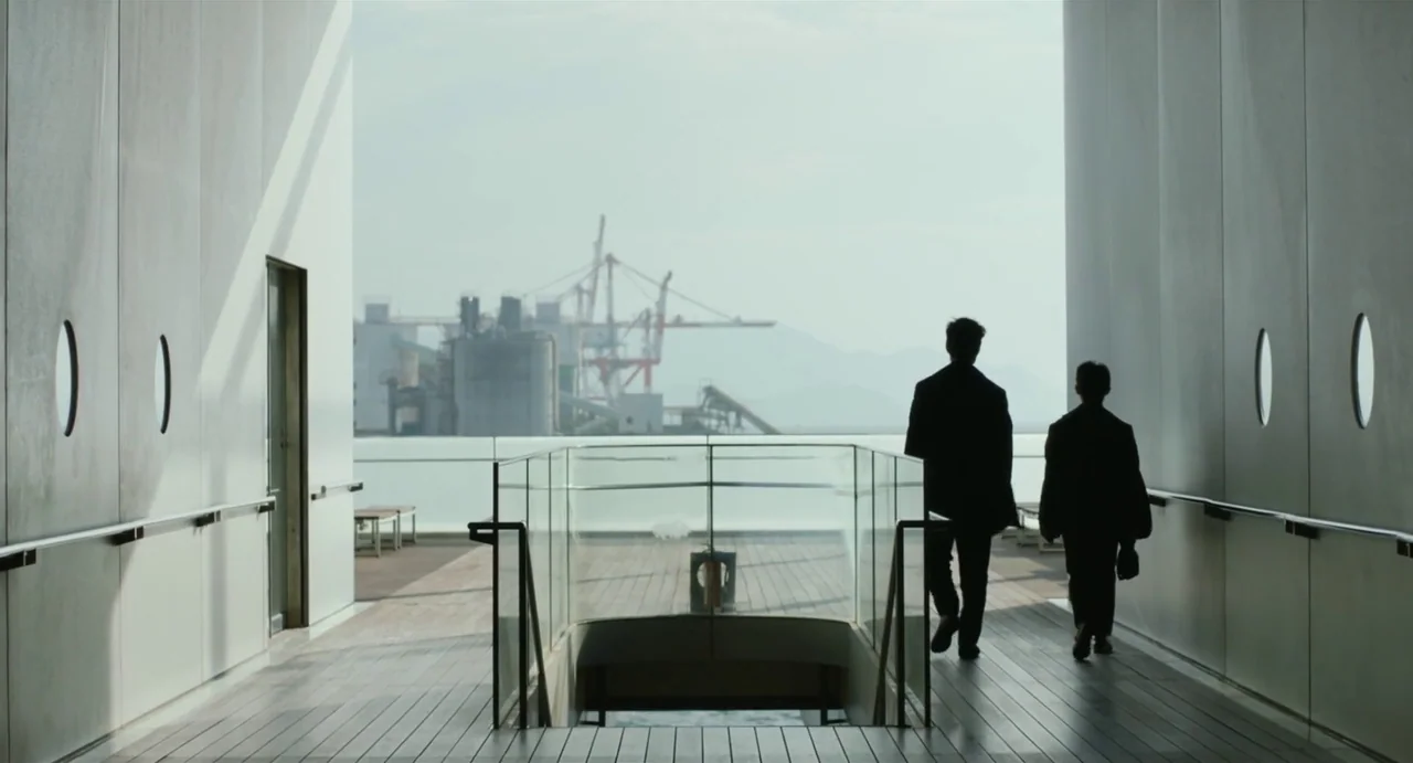 Drive My Car (2021) movie still: fullbody — An outdoor observation deck with modern railings and a view of an industrial port. Two men in dark suits…