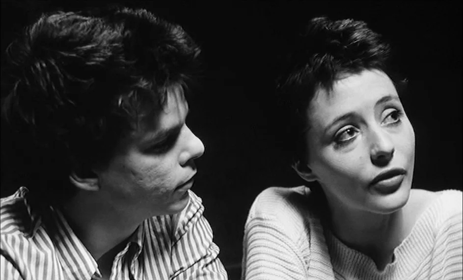 Boy Meets Girl (1984) movie still: medium — Two people are positioned closely together against an entirely dark, indistinct black background. The…