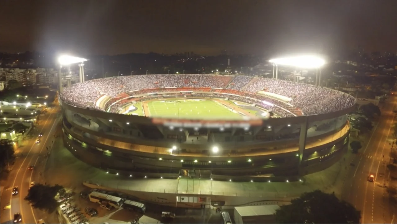 The FIFA Family: A Love Story (2017) movie still: establishing — A large illuminated football stadium stands at night surrounded by dark city…