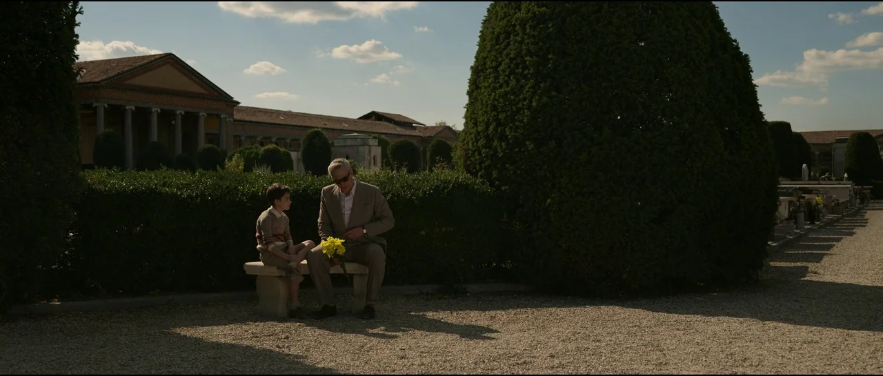 Ferrari (2023) movie still: wide — A man and a child sit on a stone bench in a cemetery. They sit before manicured hedges with a large classical…