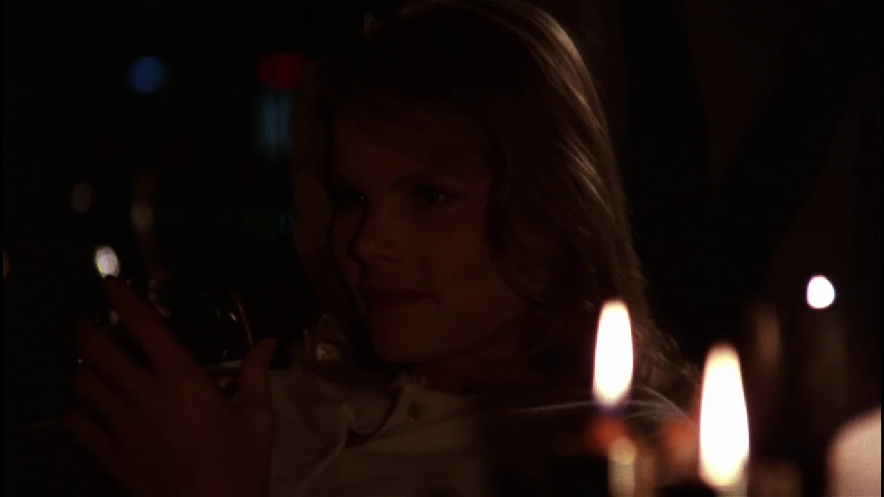 Star 80 (1983) movie still: closeup — A dark, dimly lit restaurant space features blurred lights in the distant background. A woman sits at a table…