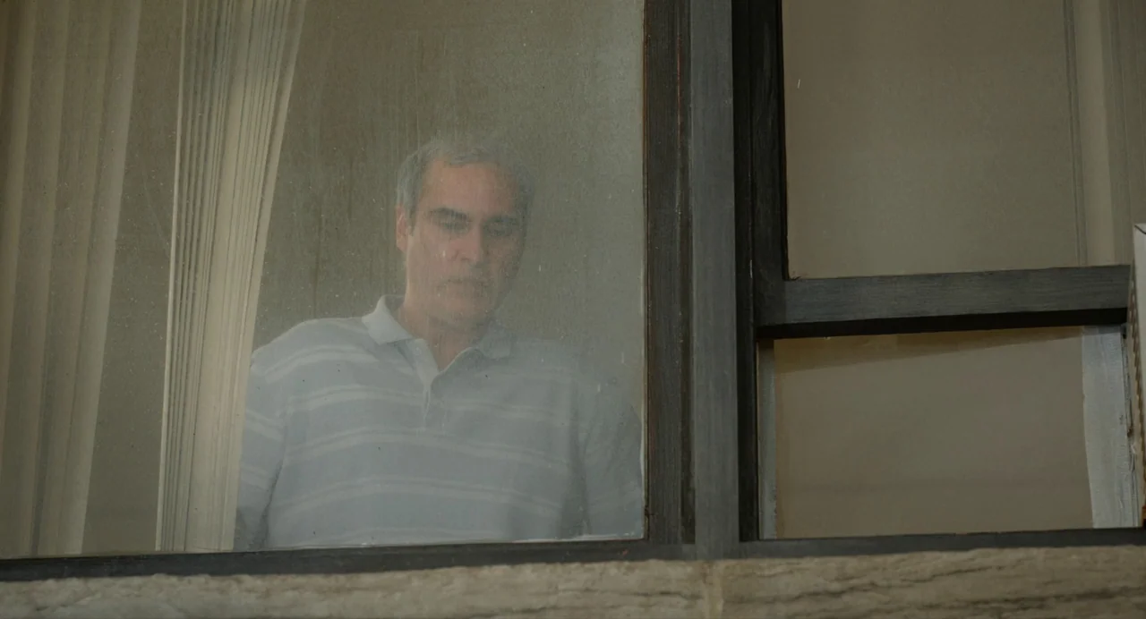 Beau Is Afraid (2023) movie still: medium — A man stands behind a slightly grimy glass window framed by dark wood. The man stares forward with a…