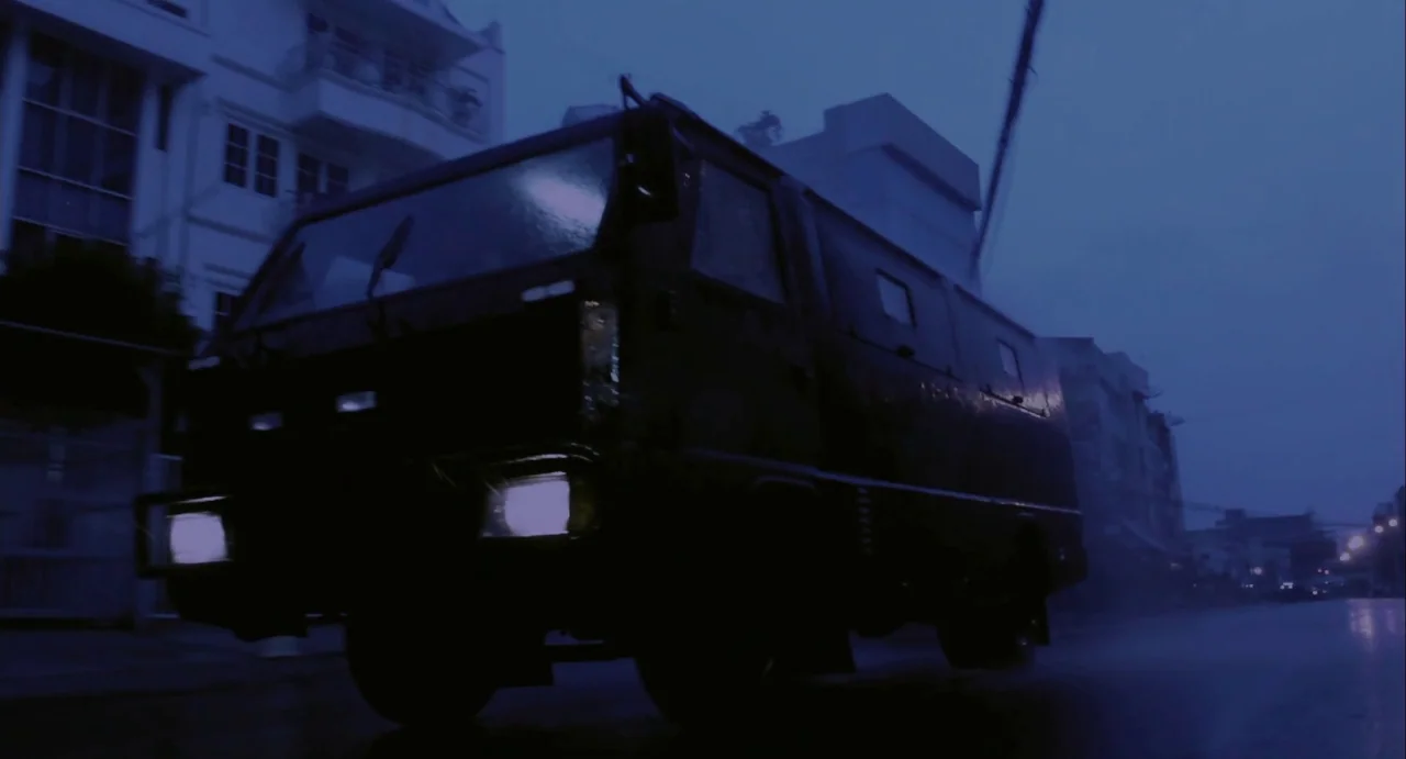 The Raid (2012) movie still: wide — An armored van drives down a wet, dark city street at night. Tall buildings flank the street in the deep blue…