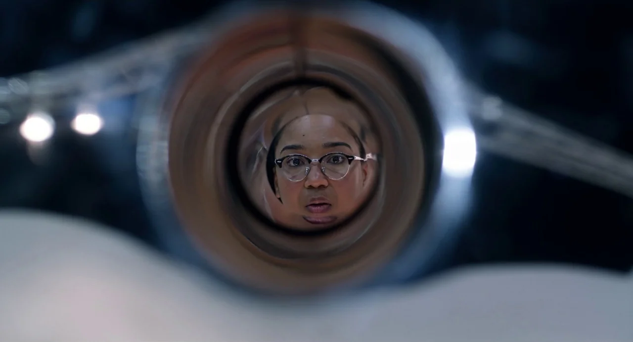 Velvet Buzzsaw (2019) movie still: closeup — A dark space focuses on a person's face reflected inside a cylindrical metal lens. The subject gazes…