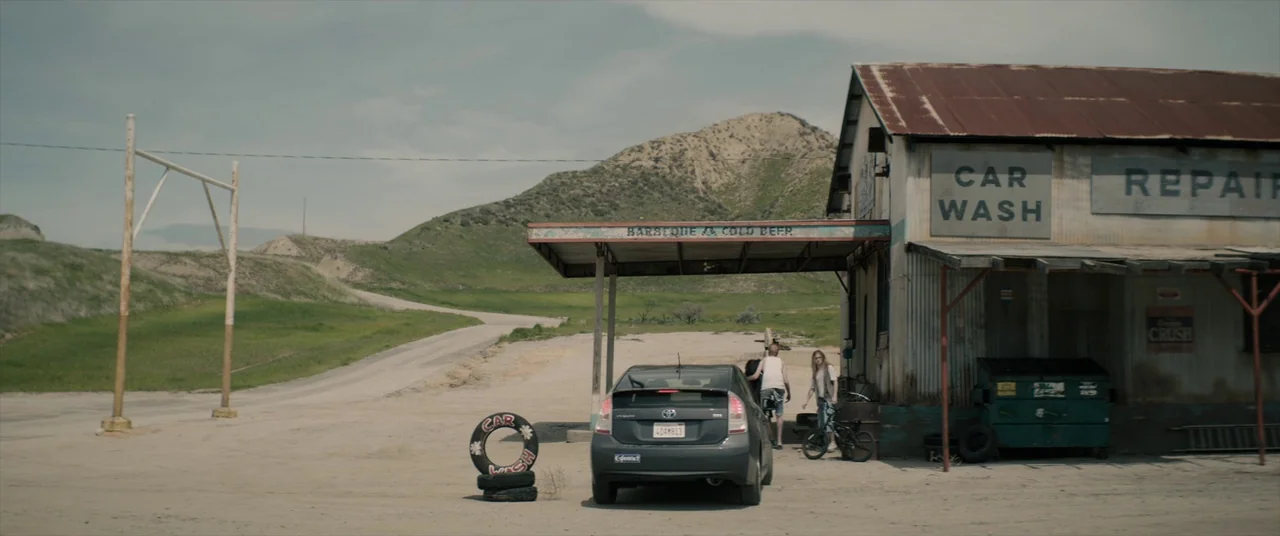 Tone Deaf (2019) movie still: wide — A rural, weathered repair shop with a rusty roof sits against a hilly landscape. Two young adults stand by a…