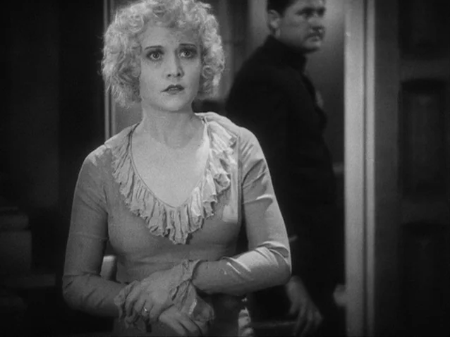 The Docks of New York (1928) movie still: medium — A dark indoor hallway with a woman standing in the foreground, clutching her hands. A man in a…