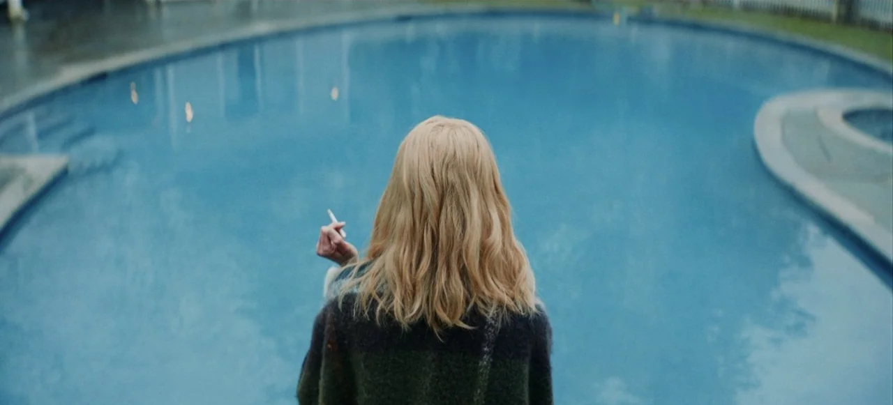 Babygirl (2024) movie still: medium — A backyard residential swimming pool with blue water dominates the frame. A woman stands in the foreground,…