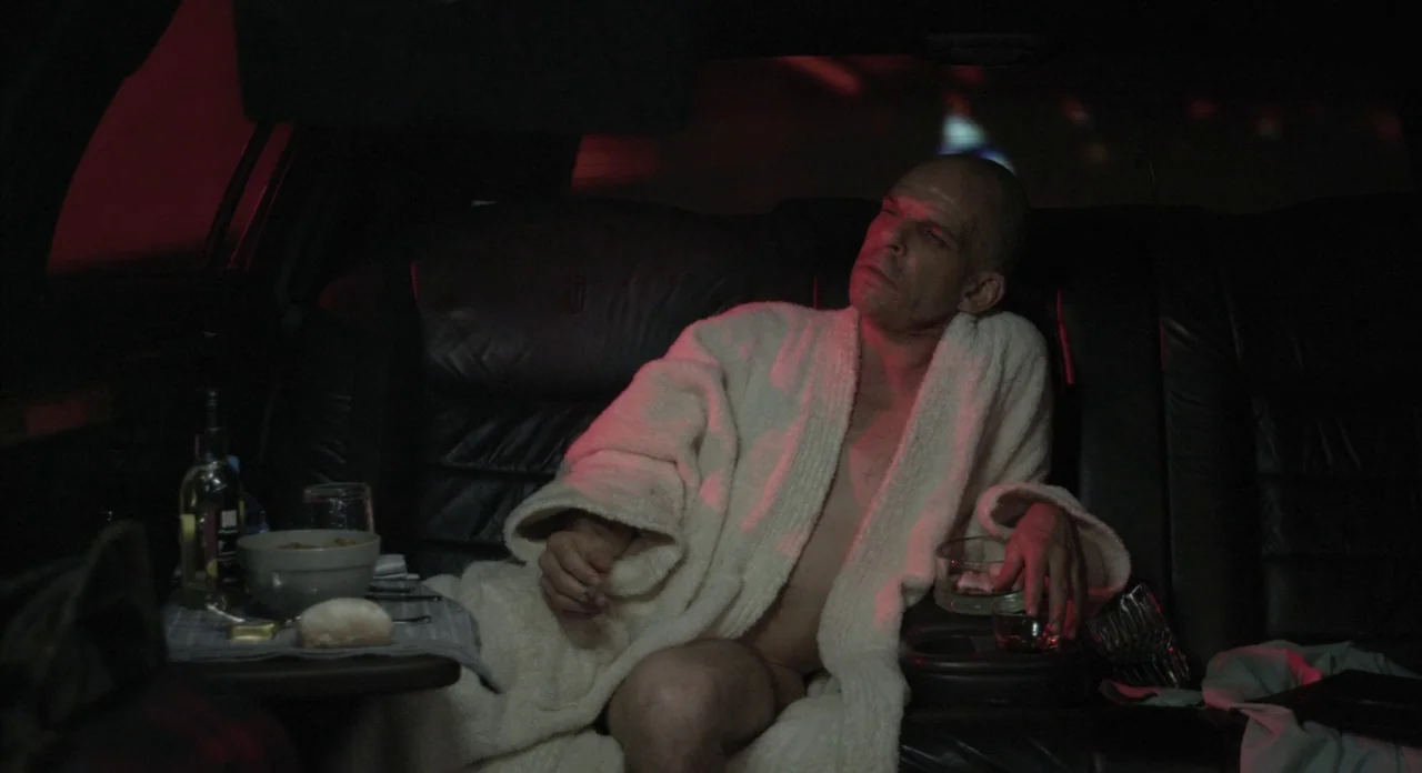 Holy Motors (2012) movie still: medium — The interior of a dark limousine features black leather upholstery and a small table. A bald man wearing a…