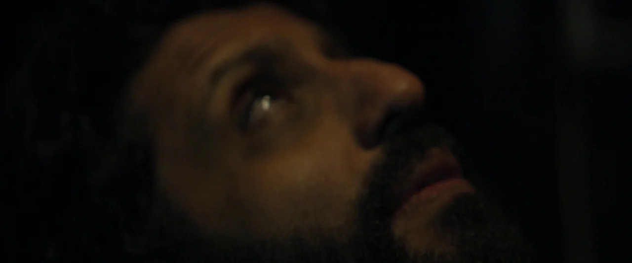 Ali & Ava (2021) movie still: closeup — A man's face emerges from deep shadow in a dark, nondescript room. He looks upward with his lips parted.