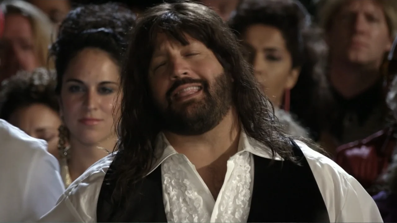 Grown Ups 2 (2013) movie still: closeup — A crowded performance space is filled with people in the dim background. A man with a dark wig and ruffled…