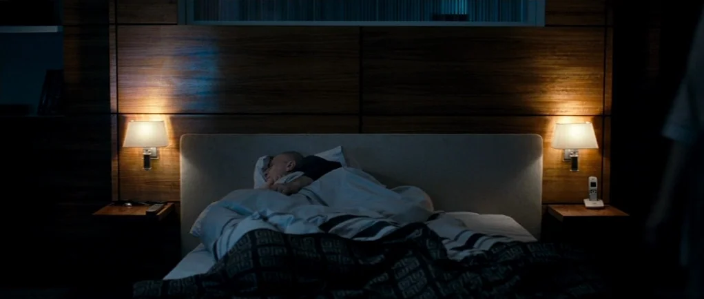 Elena (2011) movie still: medium — A modern hotel room features a horizontal wood-paneled headboard with two mounted lamps. A man lies asleep under…