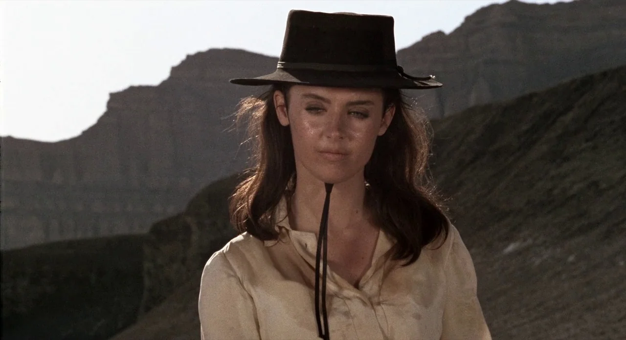 The Shooting (1966) movie still: medium — A woman stands in a vast, sun-drenched desert landscape with distant rocky formations. She wears a…