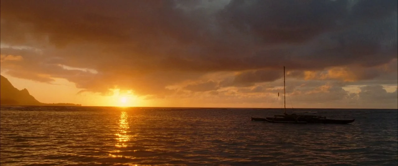 The Descendants (2011) movie still: wide — A calm expanse of water sits under a dense, golden-lit sunset sky. A lone outrigger boat is silhouetted…