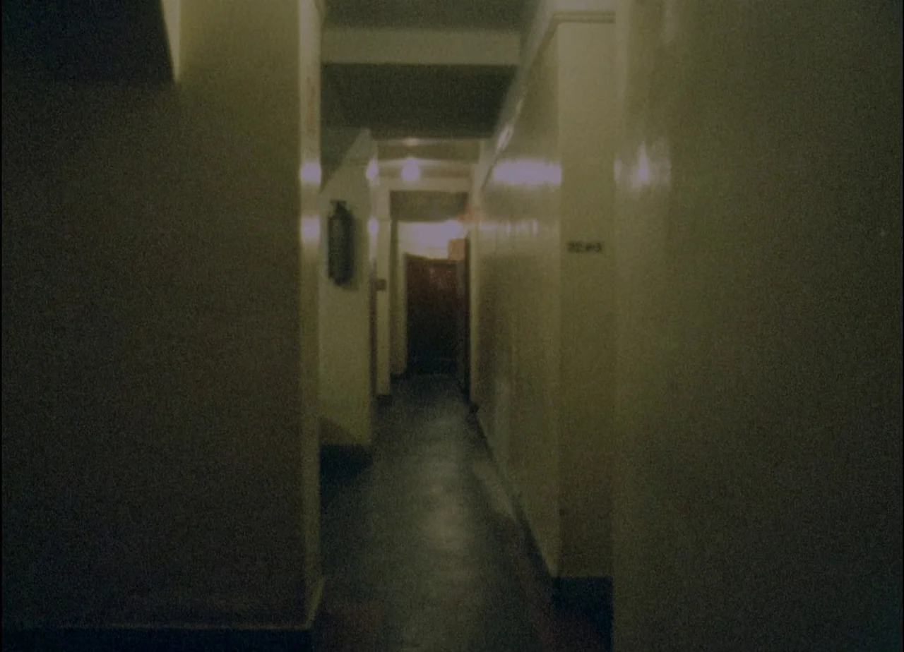 Hotel Monterey (1973) movie still: establishing — A dimly lit residential corridor features beige walls and a dark concrete floor. Light sources are…