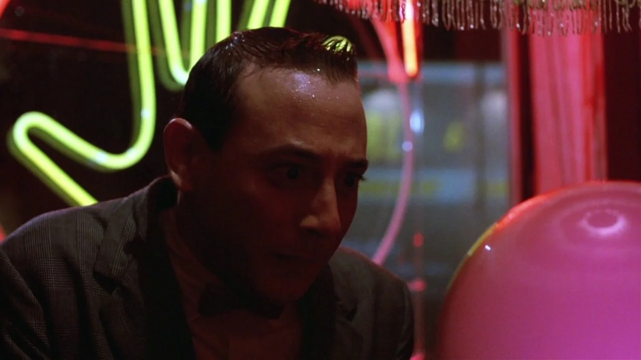 Pee-Wee’s Big Adventure (1985) movie still: closeup — An interior space lit by glowing pink and neon green signage at night. A man sits in the…