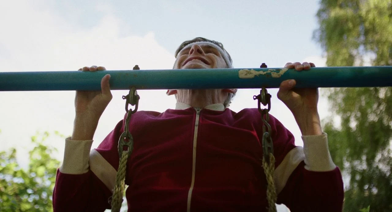 Dating Amber movie still: medium — An outdoor playground features a weathered blue metal horizontal bar against a bright sky. An older man in a…