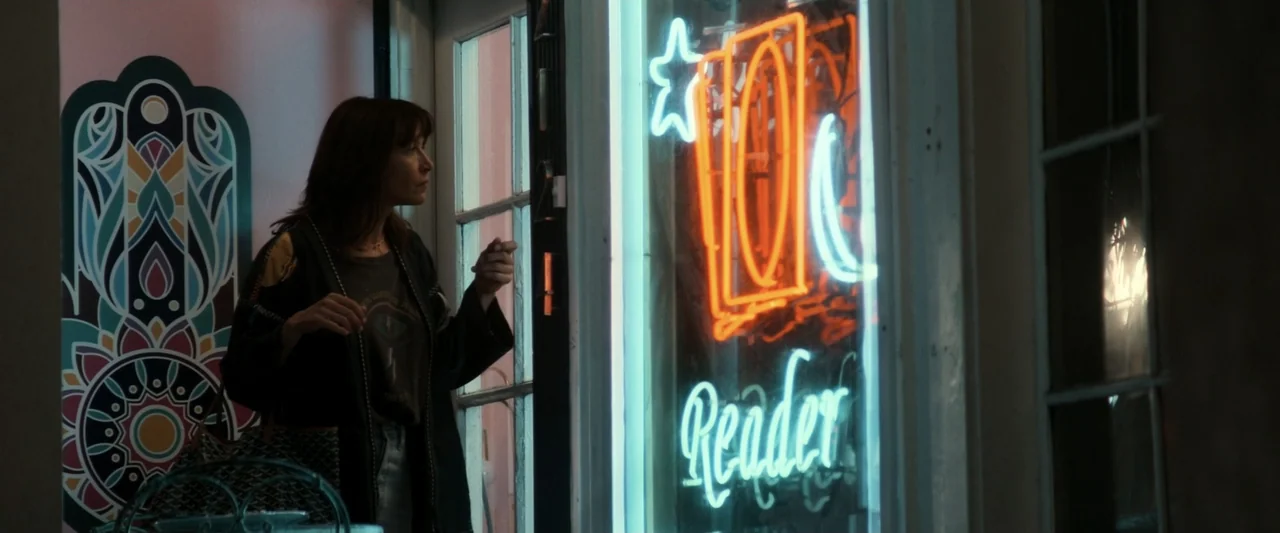 I Love America (2022) movie still: medium — A dimly lit interior space with a window featuring a bright glowing neon sign. A woman in a jacket…