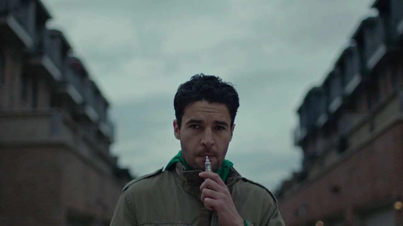 Possessor (2020) movie still: medium — A man stands in the middle of a residential street between two low-rise buildings. He holds a small…