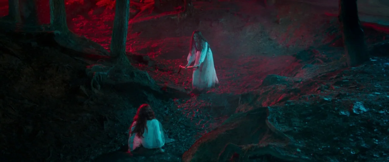 Censor (2021) movie still: wide — A dark, uneven woodland area features tree roots protruding from the muddy earth floor. One woman in a white dress…