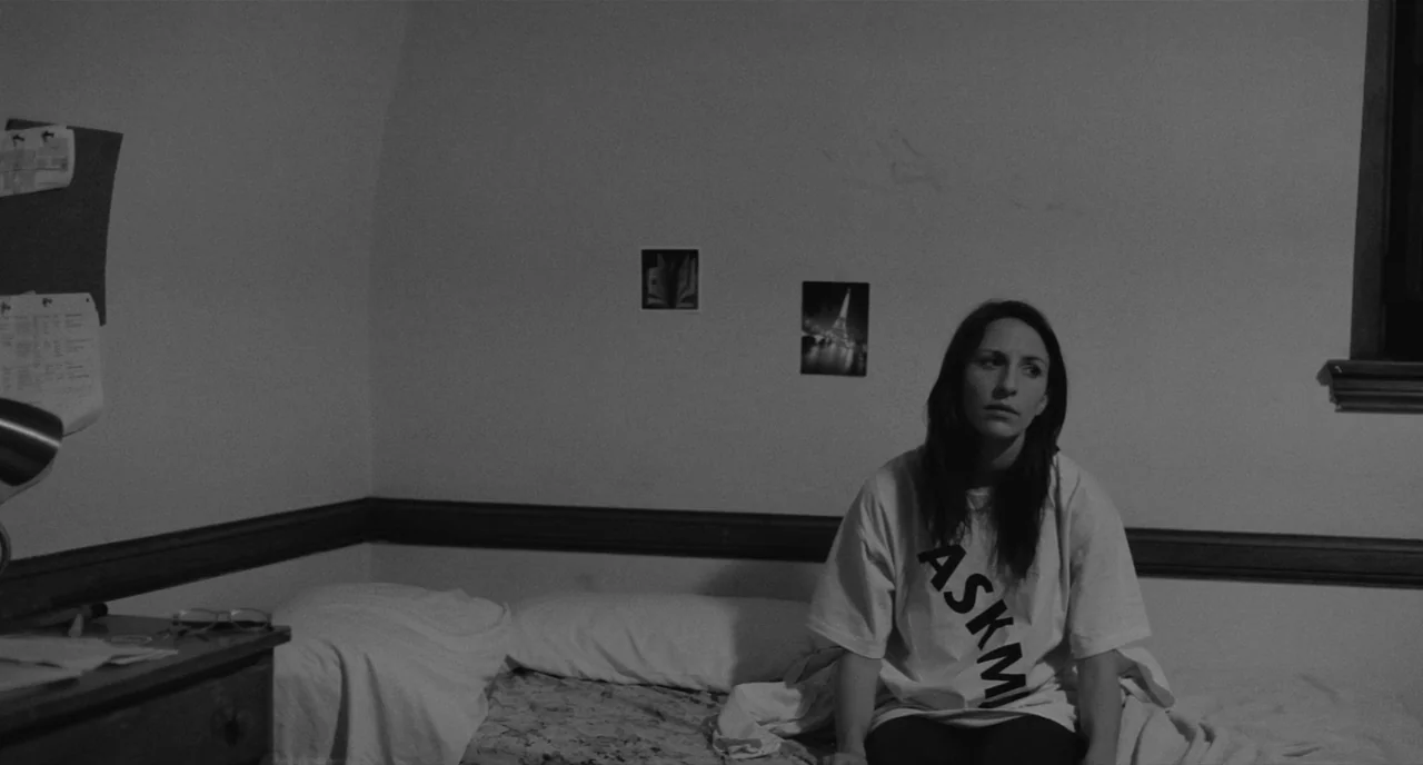 Frances Ha (2012) movie still: medium — A plain bedroom with white walls and minimal decor including two small wall prints. A woman sits on the edge…
