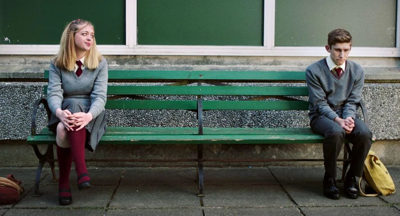 Dating Amber movie still: fullbody — A green wooden bench sits in front of a school building with large windows. A boy and girl in matching school…