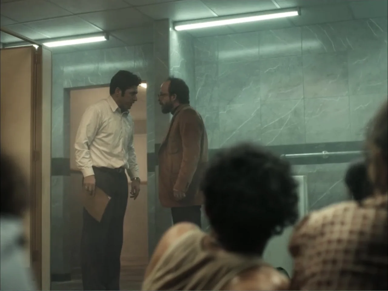 November (2025) movie still: medium — A public restroom with tiled walls and fluorescent ceiling lights. Two men stand in the center talking while…