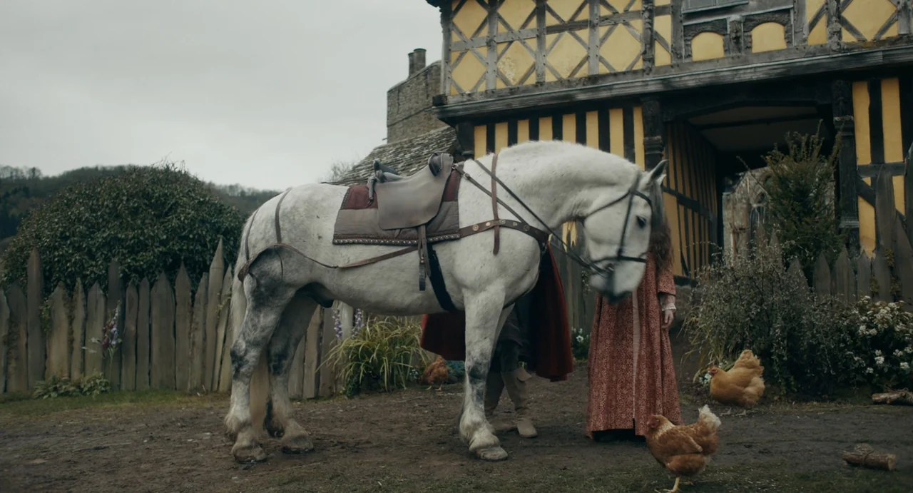 Catherine Called Birdy (2022) movie still: medium — A timber-framed medieval building stands behind a dirt courtyard with a wooden fence. A white…
