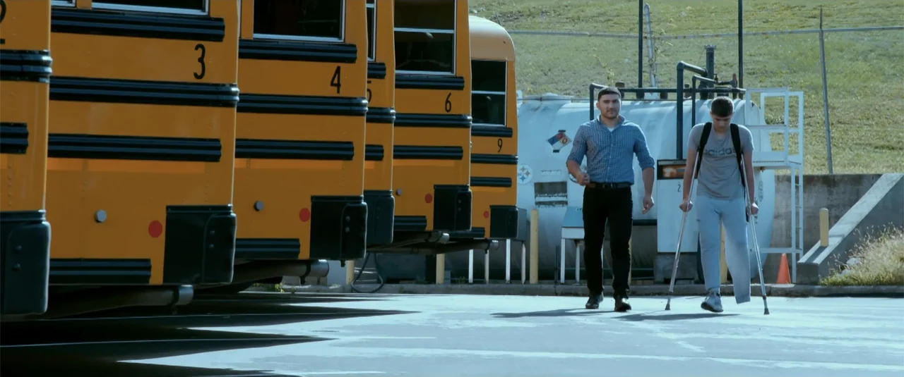 Simple as Water (2021) movie still: fullbody — A row of yellow school buses is parked on a paved lot beside an industrial white tank. Two men walk…