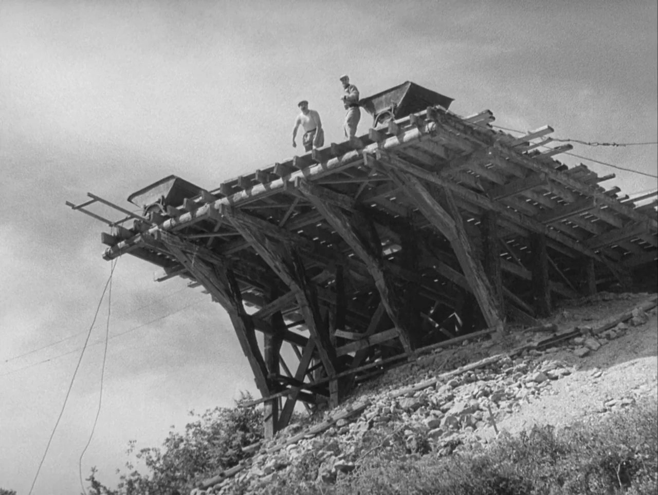 The Wages of Fear (1953) movie still: wide — A wooden temporary scaffolding structure rests on a rocky incline against a grey sky. Two male workers…