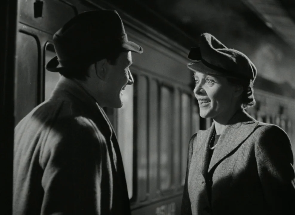 Brief Encounter (1945) movie still: medium — A dark, moody train station platform at night with a large passenger carriage behind them. A man in a…