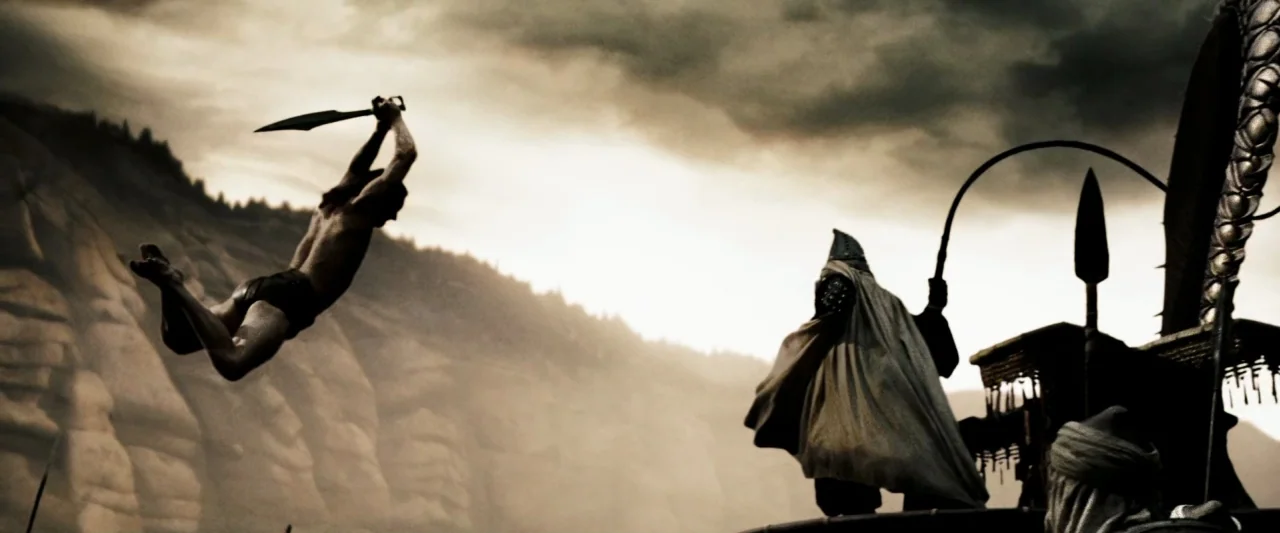 300 (2006) movie still: wide — A craggy mountain pass features sheer rock walls under a brooding, overcast sky. A shirtless warrior mid-leap strikes…