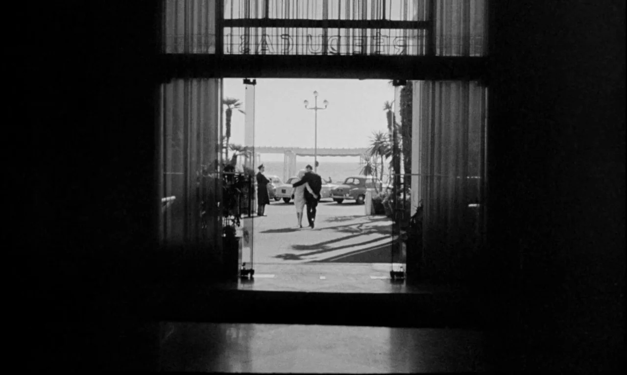 Bay of Angels (1963) movie still: wide — A dark hotel interior looks out through glass doors toward a sunny outdoor driveway. A couple walks away…