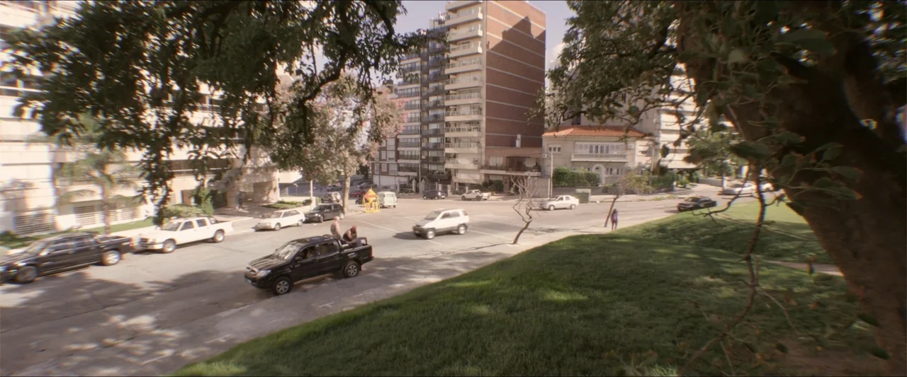 High Five (2004) movie still: wide — A street intersection borders a grassy park area with multi-story apartment buildings in background. Several…