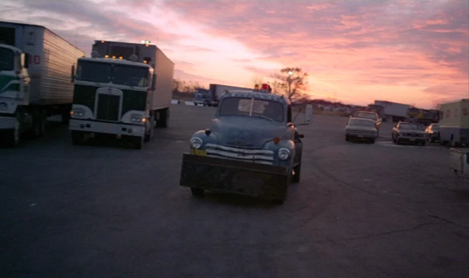 Stroszek (1977) movie still: wide — A spacious truck stop parking lot at dusk under a pink-streaked horizon. Several parked semi-trucks and a blue…
