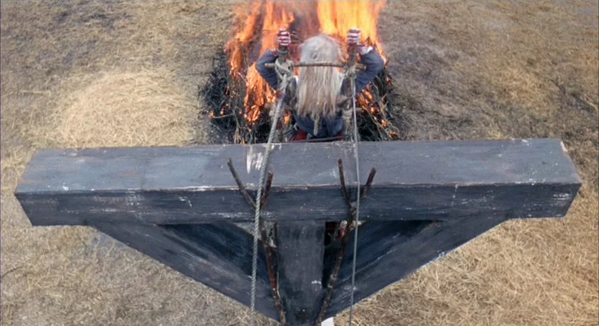 Witchfinder General (1968) movie still: wide — An outdoor area features dry straw on the ground and a large fire burning. A person stands with arms…