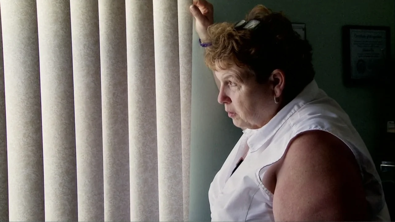 12th & Delaware (2010) movie still: medium — A woman stands indoors next to large vertical blinds covering a window. She touches the fabric of the…